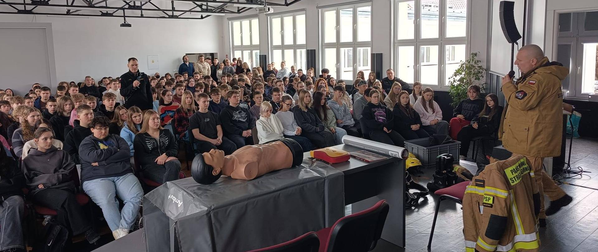 Duża sala w szkole, na krzesełkach siedzi kilkadziesiąt uczniów. Strażak z prawej strony zdjęcia przemawia do młodzieży.