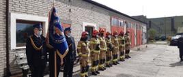 Zdjęcie przedstawia poczet sztandarowy i strażaków ze zmiany służbowej ustawionych w jednej linii na tle bram bocznych od garażu.