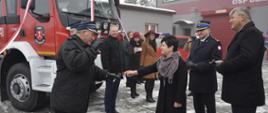 Na pierwszym planie Pani Poseł wita się z gospodarzami uroczystości. Stoi w towarzystwie komendanta wojewódzkiego PSP oraz wice wojewody kujawsko-pomoskiego. W tle fragment pojazdu z dekoracjami oraz część budynku remizy.