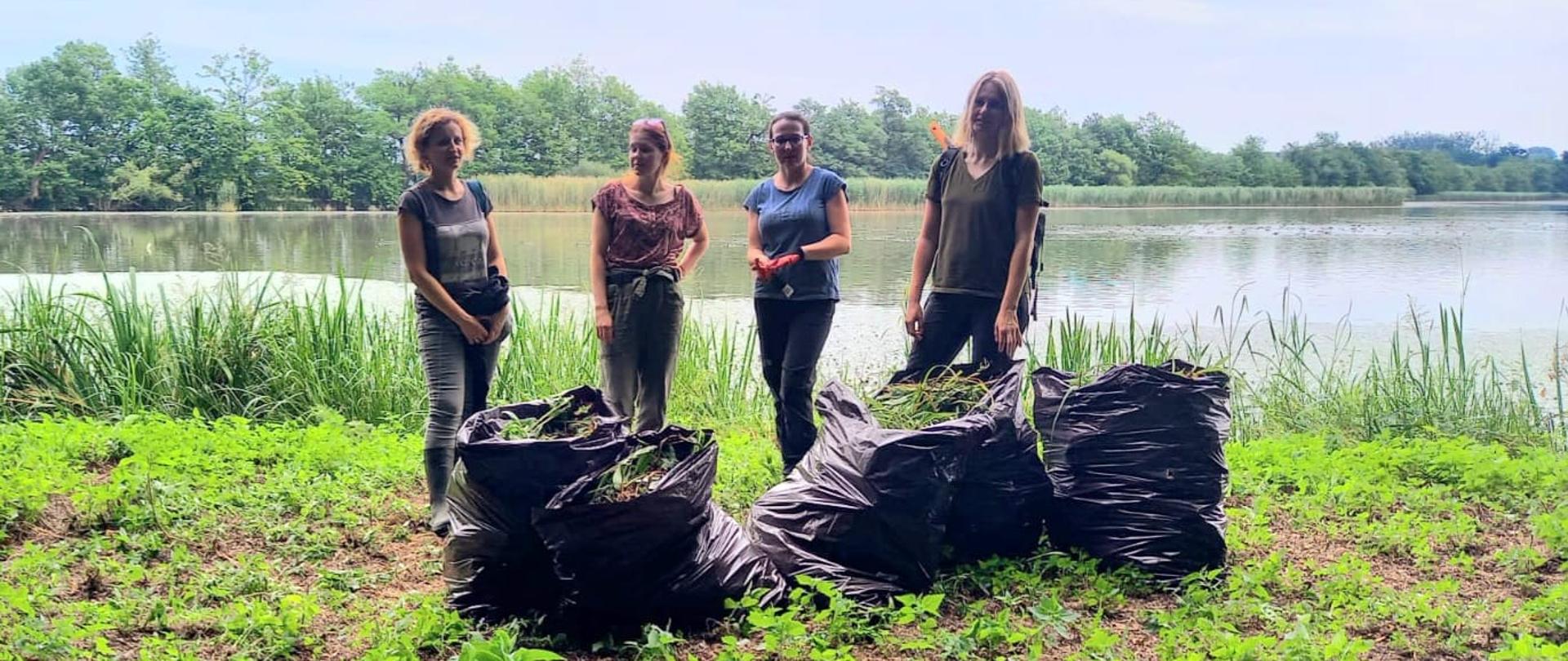 Cztery kobiety stojące na tle stawu, przed nimi worki wypełnione roślinami