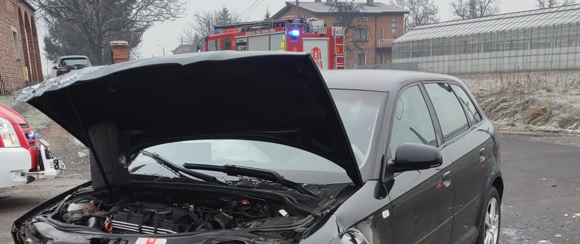 Zdjęcie na drodze publicznej. Wykonane w ciągu dnia. Zachmurzenie duże. Na drodze rozbite pojazdy oraz pojazdy straży pożarnej. Samochody ratowniczo-gaśnicze w kolorze czerwonym, posiadają sygnalizację ostrzegawczą. Widać zabudowania jednorodzinne.