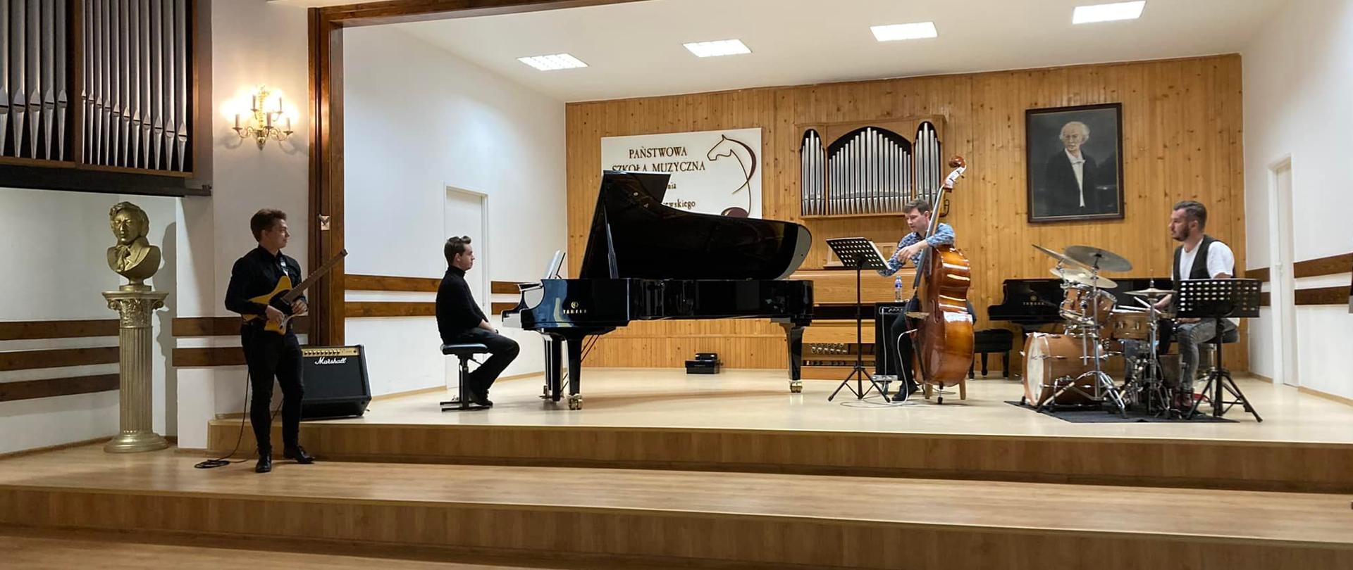 Na scenie stoi gitarzysta, z tyłu sekcja arytmiczna: fortepian, kontrabas i perkusja.