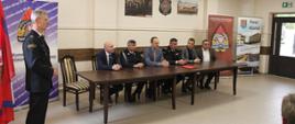 strażak w mundurze wyjściowym przedstawia gości siedzących przy stole prezydialnym