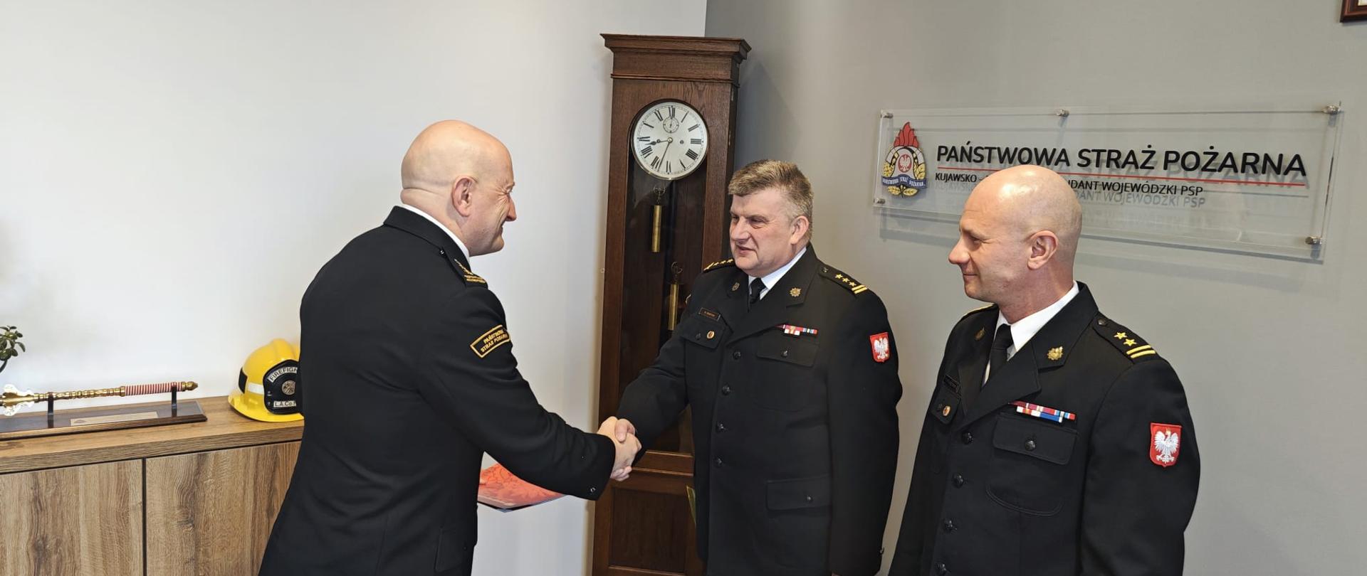 Trzech oficerów pożarnictwa. Dwóch z nich podają sobie dłonie, trzeci stoi obok. W tle na ścianie logo i napis firmowy, za nimi zegar stojący.