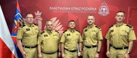Pięciu mężczyzn w beżowych mundurach Państwowej Straży Pożarnej stoi na tle czerwonej ściany z napisem "Państwowa Straż Pożarna Komenda Miejska Państwowej Straży Pożarnej w Szczecinie" i symbolem orła. Po lewej stronie kadru widoczne są dwie flagi: biało-czerwona flaga Polski oraz niebiesko-czerwona flaga z gryfem. Wszyscy mężczyźni stoją w szeregu, na oficjalnym tle z herbem i logotypem PSP.
