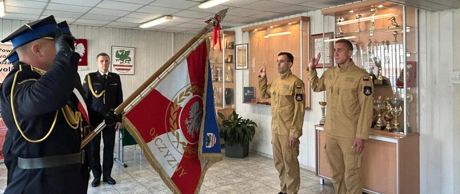 Zdjęcie przedstawia uroczystość w pomieszczeniu. Na pierwszym planie, po lewej stronie, widać mężczyznę w granatowym mundurze, który salutuje, a obok niego stoi mężczyzna trzymający sztandar z polskim godłem. Po prawej stronie zdjęcia dwóch mężczyzn w beżowych mundurach stoi na baczność, z ręką podniesioną do góry, składając przysięgę.