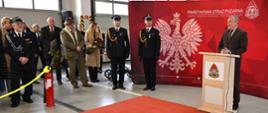 Poseł przemawia przy mównicy na tle ścianki z boku strażacy oraz inne osoby