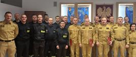 Na zdjęciu znajduje się grupa funkcjonariuszy Państwowej Straży Pożarnej pozujących do wspólnej fotografii po zakończeniu uroczystości służbowej. Wśród uczestników widoczni są strażacy w umundurowaniu służbowym i specjalnym, a część z nich trzyma czerwone teczki związane z otrzymanymi wyróżnieniami lub awansami. W tle znajdują się sztandary i symbole związane ze strażą pożarną oraz godło Rzeczypospolitej Polskiej.