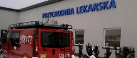 Na zdjęciu widać osobowy samochód strażacki o numerach operacyjnych 508R73 na tle budynku z dużym niebieskim napisem „PRZYCHODNIA LEKARSKA” na elewacji. Wkoło widać zalegający śnieg