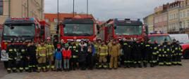 Zdjęcie grupowe wykonane na rynku. Na pierwszym planie widać strażaków oraz wolontariuszy, za nimi ustawione są pojazdy ratownicze. W tle widać ratusz miejski oraz inne zabudowania.