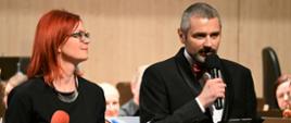 Zdjęcie kolorowe przedstawiające konferansjerów koncertu w filharmonii stojących na scenie. W tle orkiestra symfoniczna.