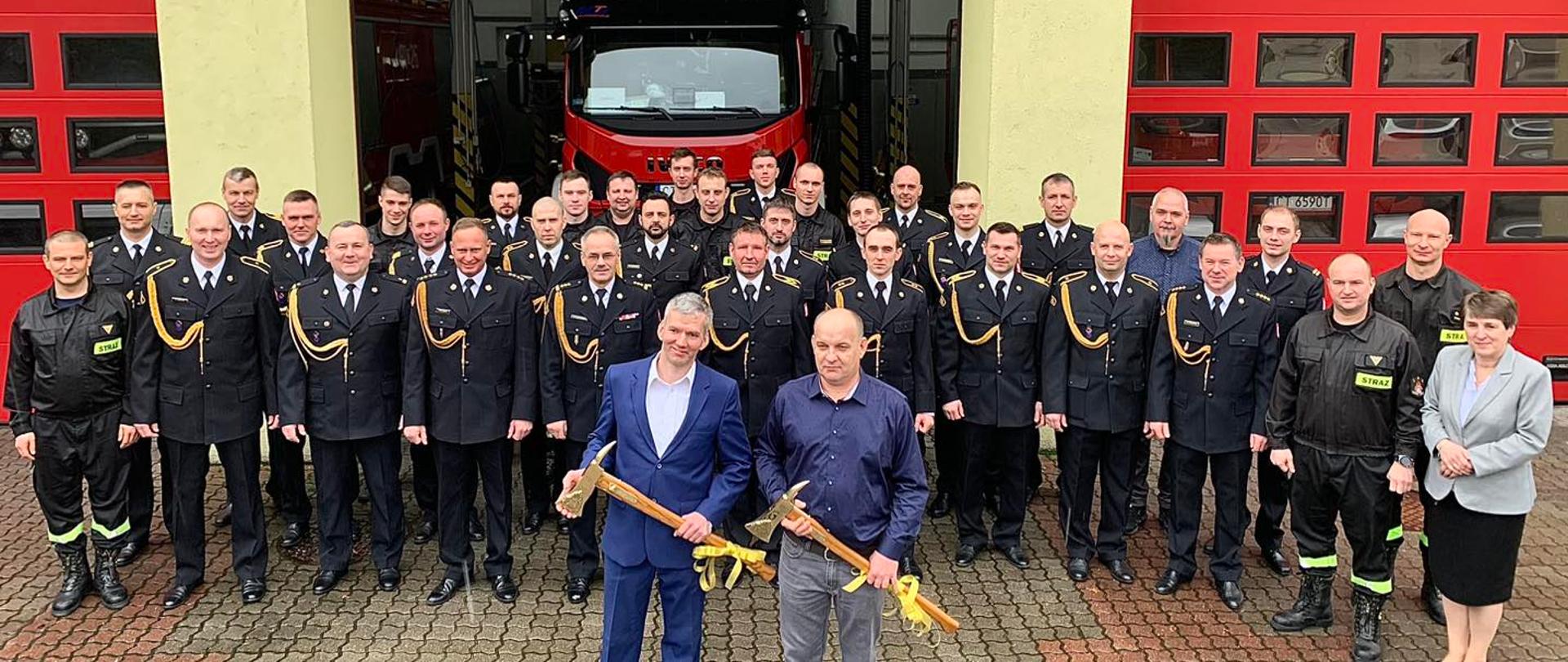 Strażacy stoją przed budynkiem komendy. Emeryci trzymają pamiątkowe topory strażackie. W tle wóz pożarniczy