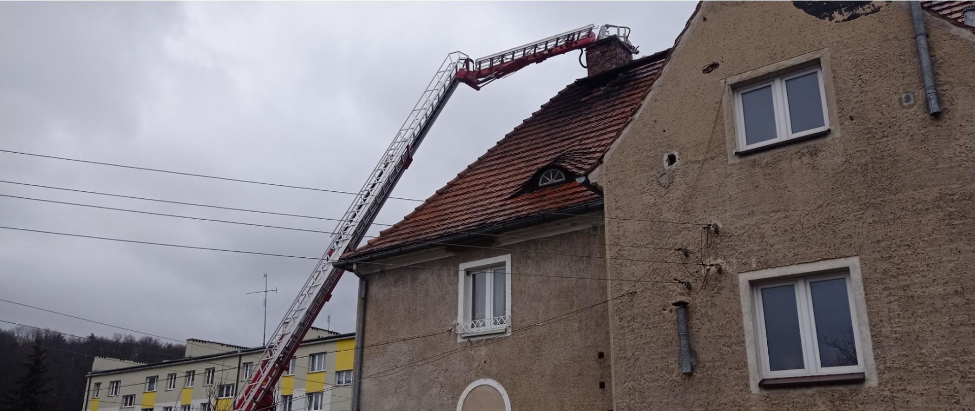 Obraz przedstawia rozstawiony samochód typu podnośnik. Kosz podnośnika znajduje się przy kominie. Na pierwszym planie budynek mieszkalny. W tle blok mieszkalny oraz las.