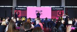 Zdjęcie od tyłu sali, wiceminister Zioło-Pużuk w stoi na tle różowo-fioletowej ściany z napisem POP Science i mówi do mikrofonu na stojaku, przed nią na sali siedzi na krzesłach dużo ludzi.