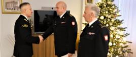 Komendant główny PSP wręcza decyzję p. o. zastepcy mazowieckiego komendanta wojewódzkiego PSP stojąc także przed mazowieckim komendantem wojewódzkim PSP w tle ubrana choinka 