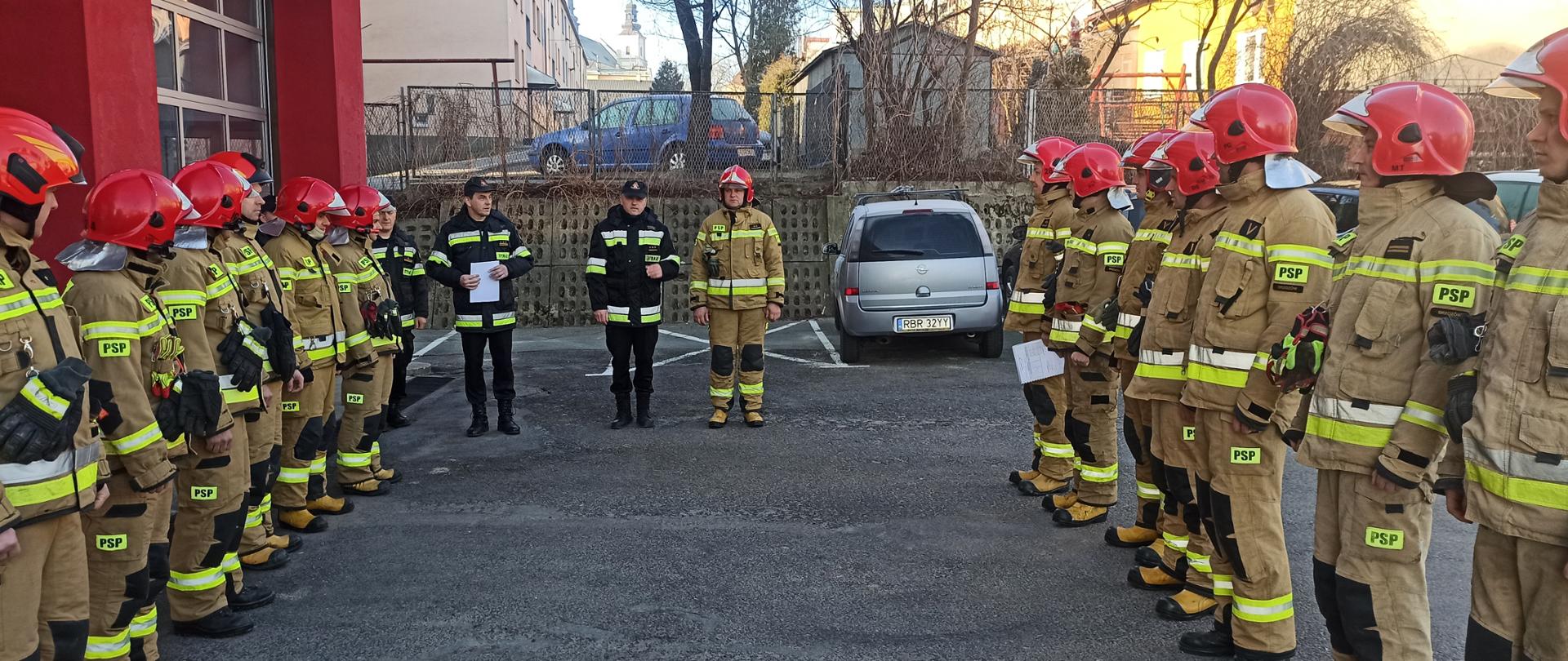 Kolorowe zdjęcie wykonane na zewnątrz, w pochmurny dzień przedstawia umundurowanych strażaków na tle bram wyjazdowych budynku straży pożarnej. Strażacy stoją na zbiórce w dwóch szeregach, naprzeciw siebie. Pośrodku stoją inni strażacy prowadzący zbiórkę.