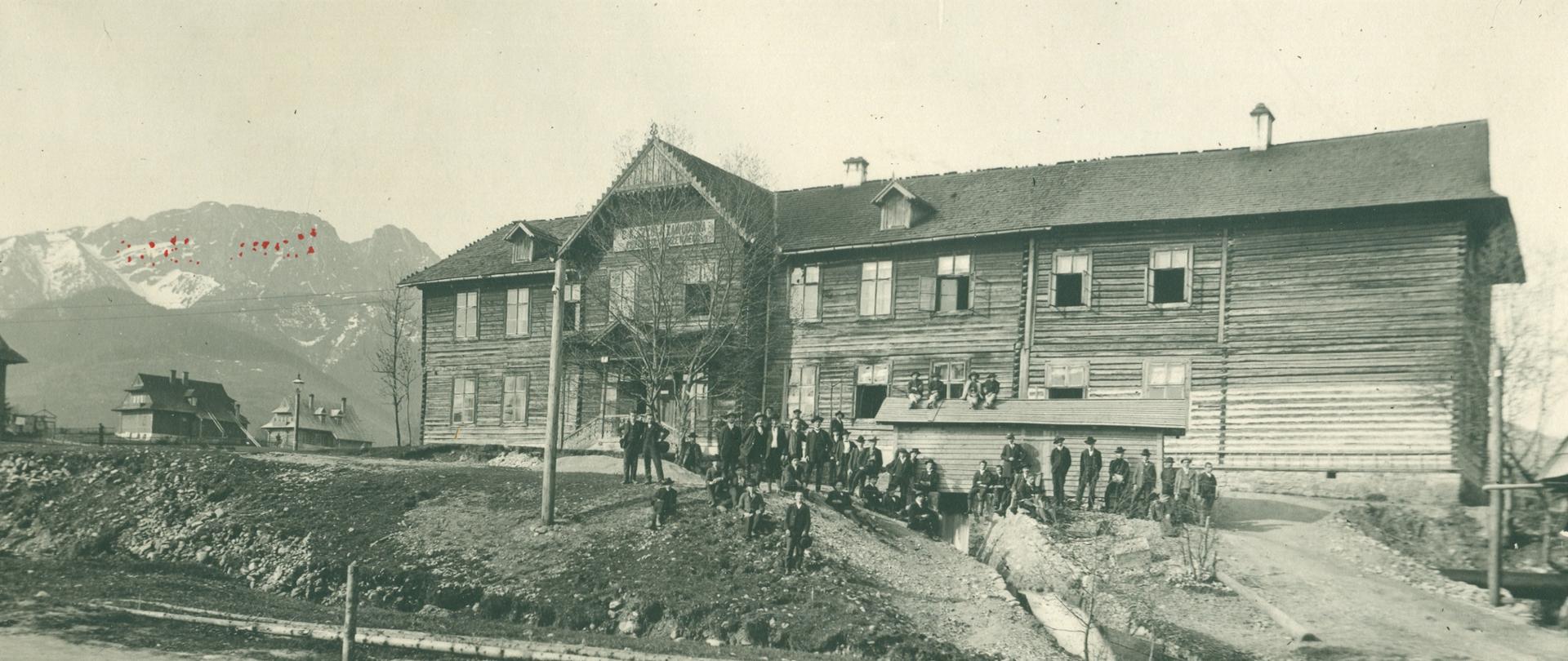 Na zdjęciu widzimy budynek Cesarsko-Królewskiej Szkoły Zawodowej Przemysłu Drzewnego. Fotografia została wykonana około roku 1900. Na pierwszym planie widzimy drogę z krawężnikiem i chodnikiem dla pieszych. Dalej widzimy koryto rzeki i skarpę, na której stoi duży drewniany budynek szkoły. Budynek zbudowany jest z poziomych belek, ma dwuspadzisty gątowy dach. W ścianach na parterze i na pierwszym piętrze osadzone są okna. Jednym z głównych elementów szkoły jest drewniany ganek - wejście do szkoły. Nad gankiem widnieje napis: C.K Szkoła Zawodowa Przemysłu Drzewnego. Przed szkoła widzimy grupę nauczycieli i uczniów. Część z nich siedzi na skarpie a część na nie stoi. Czwórka uczniów w strojach góralskich siedzi na daszku wybudowanej przed szkołą szopy na drewno.