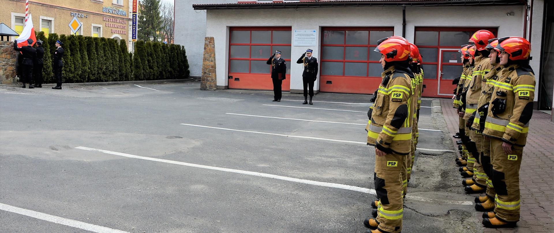 Strażacy stojący w dwuszeregu, w oddali poczet flagowy z flagą rzeczypospolitej polskiej oraz oddający honor Komendant Powiatowy wraz z dowódcą uroczystości