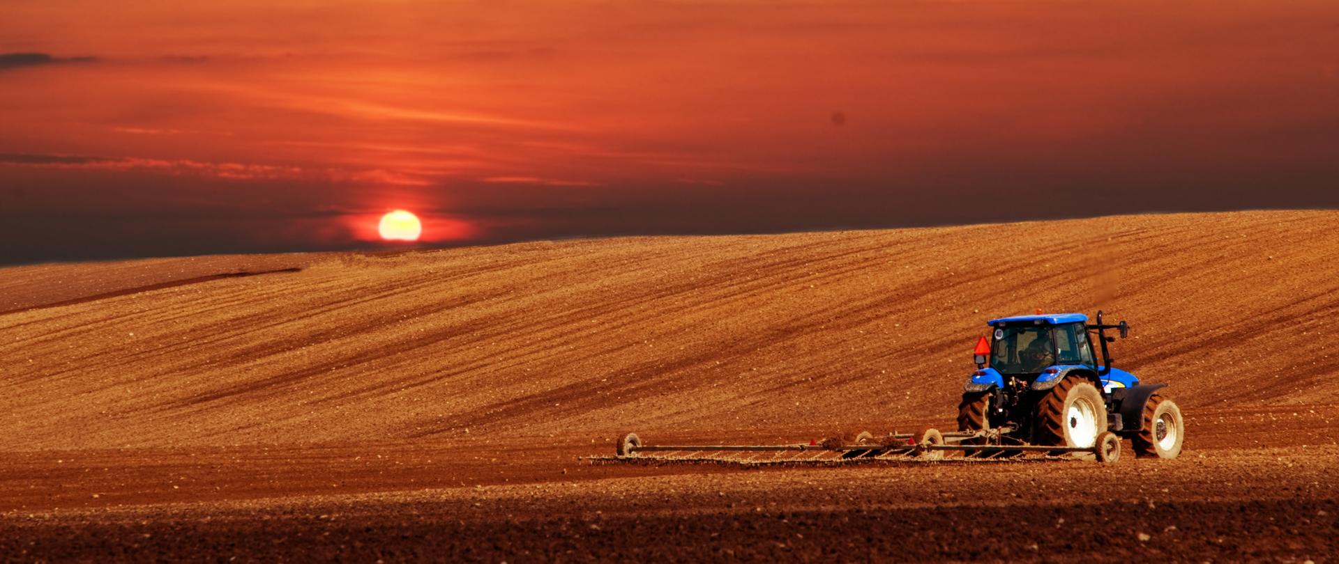 Zdjęcie przedstawia pejzaż pola podczas zachodu słońca, na polu widać traktor, który orze ziemię.
