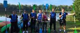Panoramiczne zdjęcie przedstawiające grupę uczniów w kamizelkach ratunkowych trzymających pionowo wiosła na trawiastym brzegu jeziora w słoneczny dzień.
