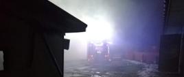 Rozwinięty, oświetlony samochód gaśniczy na tle posesji przed budynkami w których miał miejsce pożar.