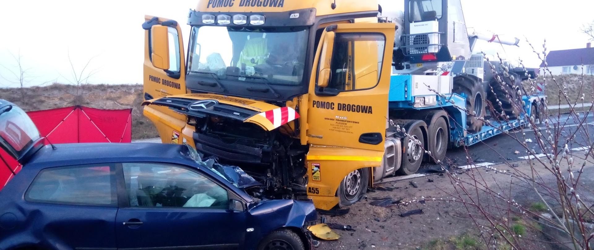 Na pierwszym planie niebieski samochód osobowy z otwartym tylnym bagażnikiem i pogniecionym przodem na skutek zderzenia z samochodem ciężarowym stojącym przy osobówce z otwartymi drzwiami pasażera i kierowcy, na naczepie przewożony pojazd, w tyle za pojazdami widoczny czerwony parawan Straży Pożarnej.