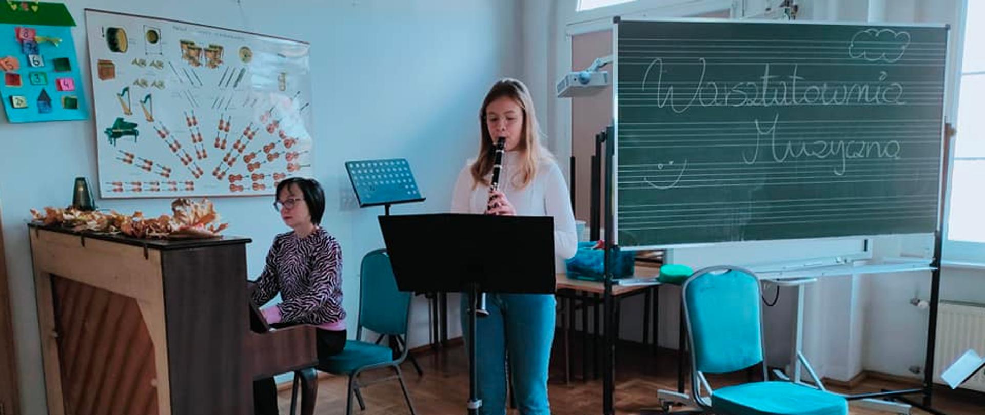 Zdjęcie przedstawia dziewczyne grającą na klarnecie w centralnej części oraz panią akompaniującą na pianinie po lewej stronie. Z prawej widnieje tablica z piećioliniami i napisam Warsztatowani muzyczna, a za nią stoją poustawiane łąwki szkolne. 