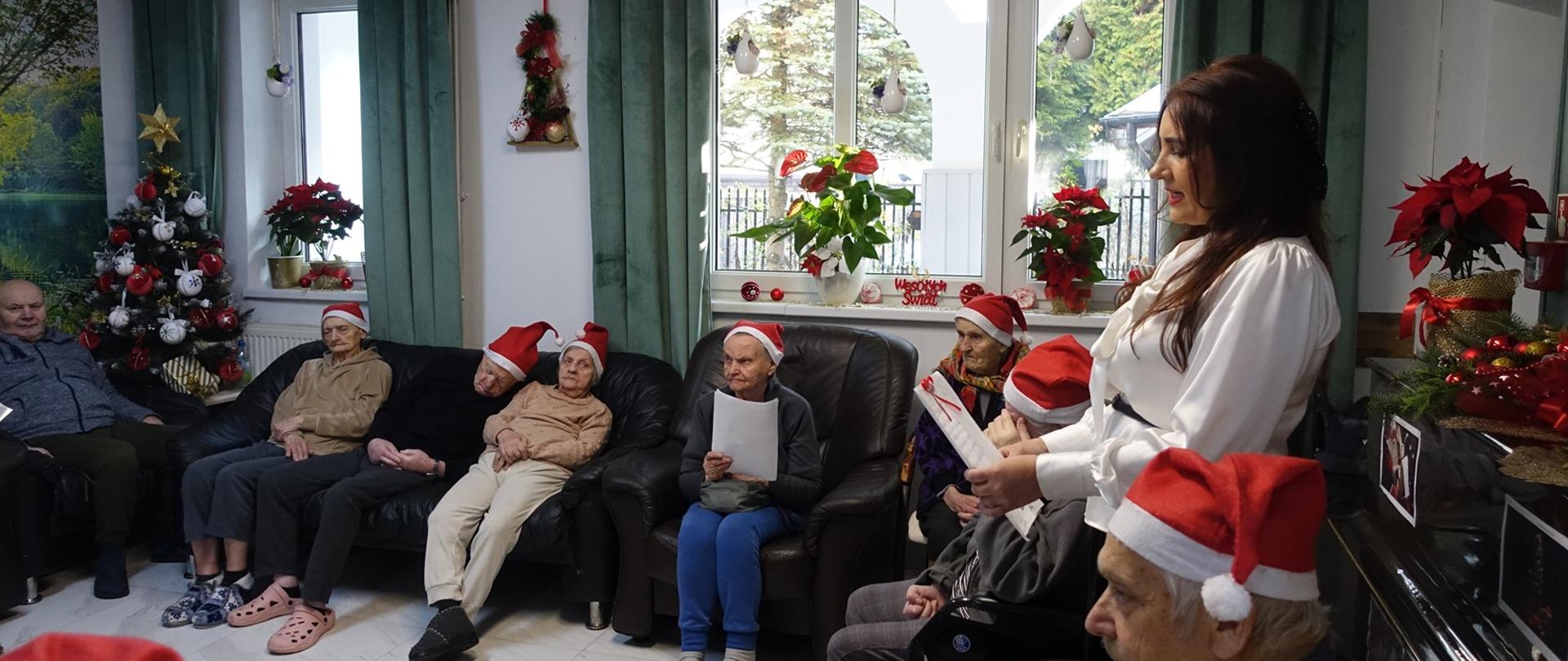  Na zdjęciu widać pensjonariuszy domu opiekuńczo-rehabilitacyjnego zgromadzonych w jasnym, przytulnym salonie. Starsze osoby siedzą na kanapach i fotelach, część ma czapki Mikołaja, co podkreśla świąteczny charakter spotkania. Obok nich stoi opiekunka, przekazująca życzenia od wszystkich podopiecznych. W tle widoczne są świąteczne dekoracje i choinka, tworzące ciepłą, domową atmosferę.