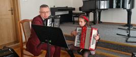 Zdjęcie przedstawia od lewej strony siedzącego mężczyznę trzymającego akordeon oraz uczennicę trzymającą akordeon i obserwującą zapis nutowy na pulpicie do nut na tle drzwi do sali i beżowej ściany.