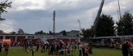 Teren Komendy Miejskiej sfotografowany w czasie trwającego Pikniku Rodzinnego gdzie na pierwszym planie widoczni przybyli goście przy stanowisku do nalewaków. w tle widoczne samochody pożarnicze oraz budynek magazynowy.