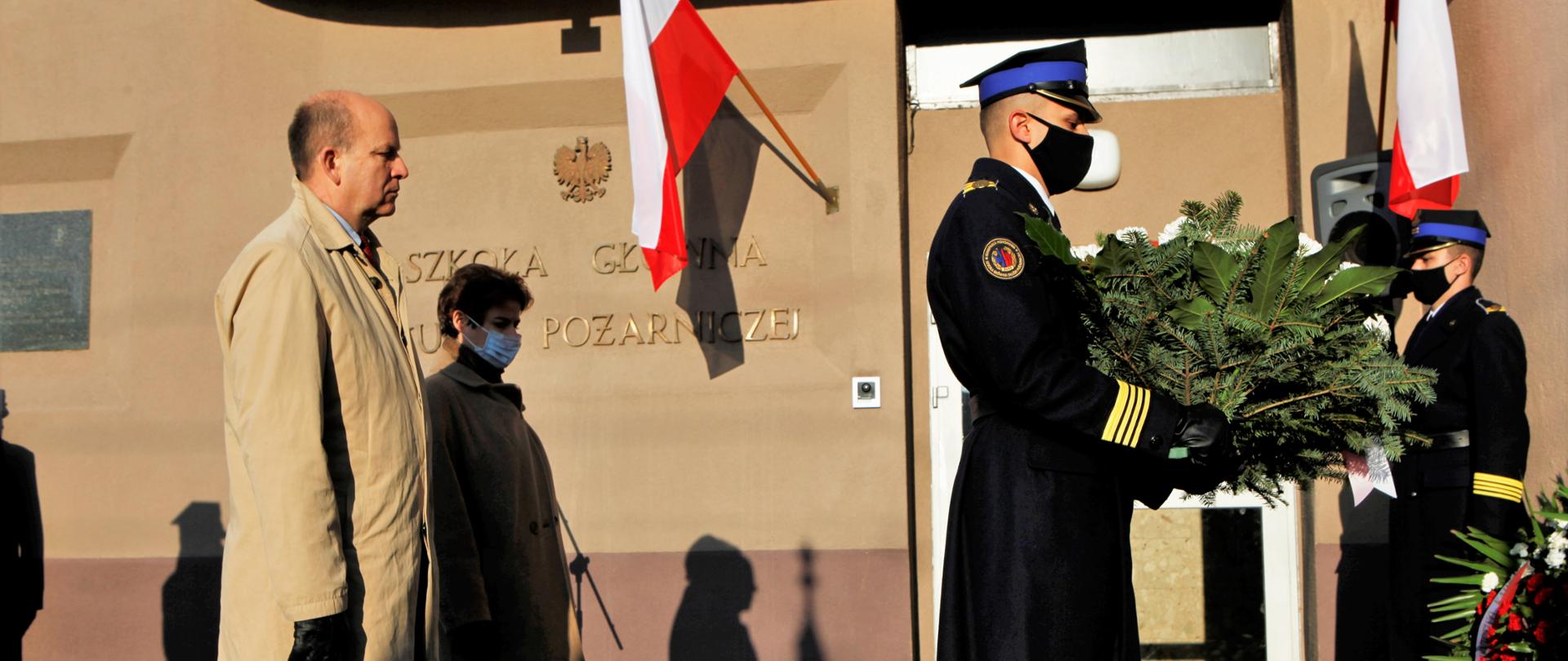 Zdjęcie przedstawia moment składania wieńców przed tablicą upamiętniającą strajk w Wyższej Oficerskiej Szkole Pożarniczej, która znajduje się przy Szkole Głównej Służby Pożarniczej w Warszawie. Na zdjęciu znajduje się strażak, asystujący w ceremonii składania wieńców. Tuż za nim znajdują się elegancko ubrani kobieta i mężczyzna.