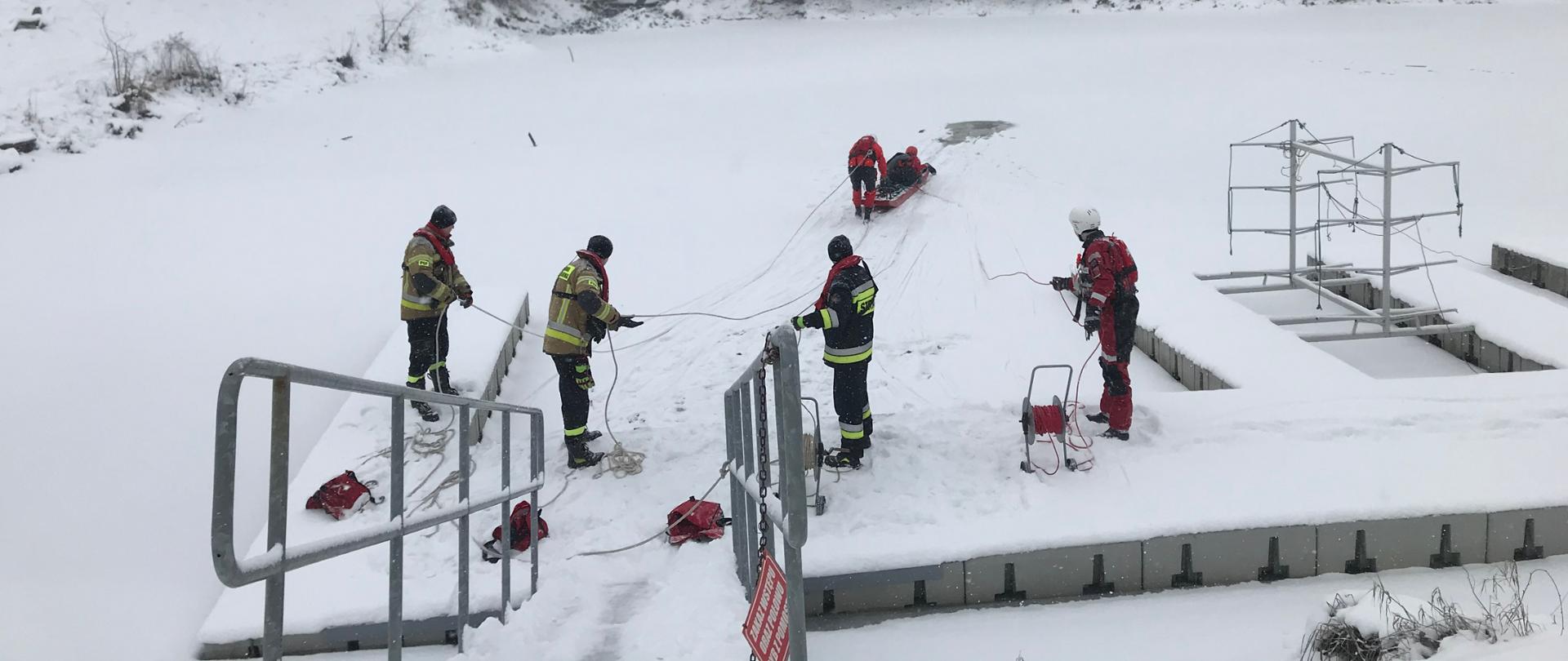 zdjęcie przedstawia zamarznięty zbiornik wodny i siedmiu strażaków. Dwóch strażaków przy pomocy sań lodowych wyciągają poszkodowanego strażaka z wody. Czterech strażaków stoi na brzegu i asekuruje przy pomocy lin strażaków, którzy są na tafli lodu i wyciągają poszkodowanego. 