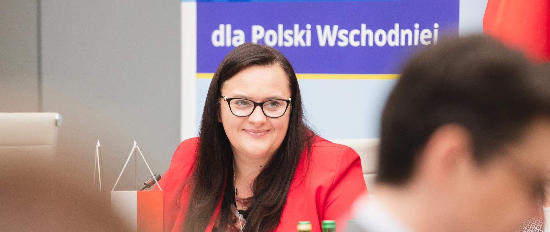 Uśmiechnięta wiceminister Małgorzata Jarosińska-Jedynak siedzi przy stole.