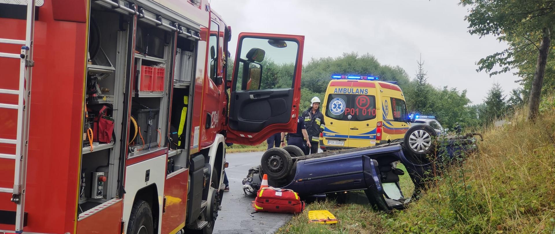 Na zdjęciu widać bok samochodu pożarniczego z otwartymi skrytkami sprzętowymi. Przed samochodem strażackim widać rozbite auto w pozycji na kołach. Dalej stoi karetka pogotowia oraz radiowóz policyjny