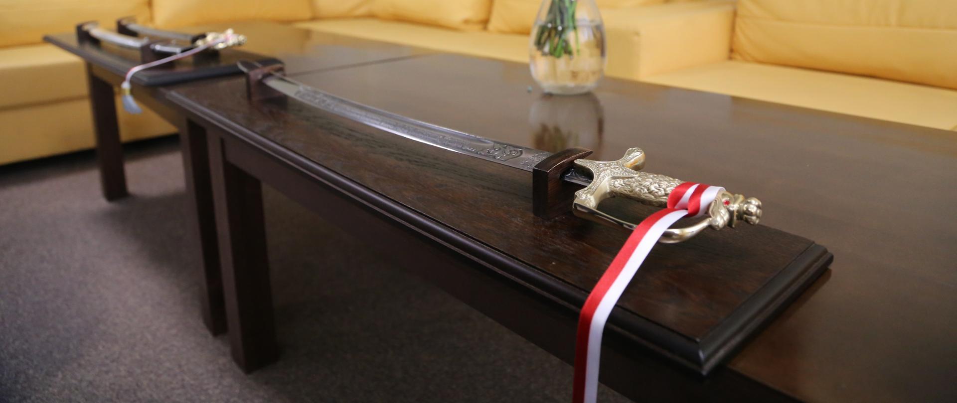 Na pierwszym planie widoczna, leżąca na ławie ozdobna szabla oficerska umieszczona w ozdobnej desce. Rękojeść szabli przepasana biało-czerwoną wstążką. Obok w tle druga szabla