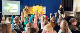 Pogadanka edukacyjna w szkole dzieci podnoszą ręce zgłaszając się do odpowiedzi. Na drugim planie strażacy.