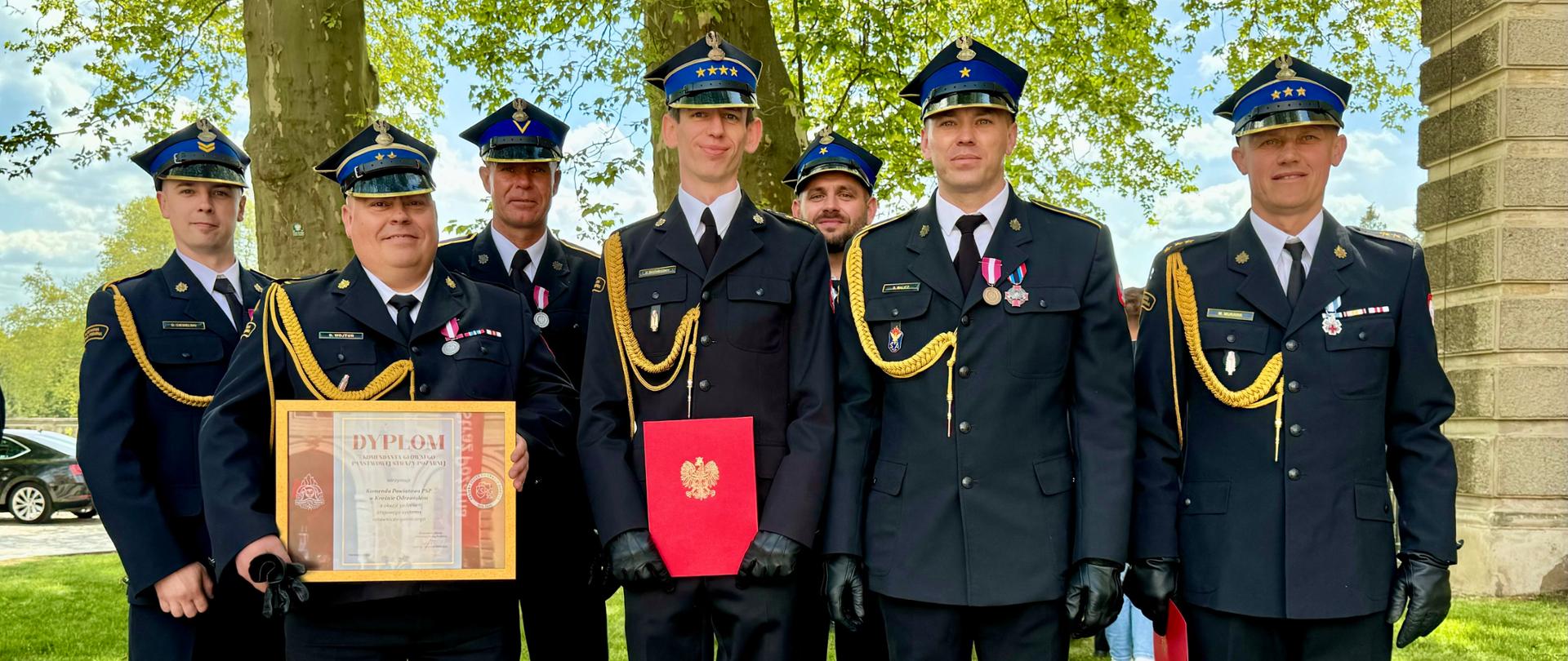 Funkcjonariusze Komendy Powiatowej Państwowej Straży Pożarnej w Krośnie Odrzańskim w mundurach wyjściowych po otrzymaniu awansów oraz odznaczeń po Wojewódzkich Obchodach Dnia Strażaka. Dwóch strażaków trzyma dyplom i czerwoną teczkę.