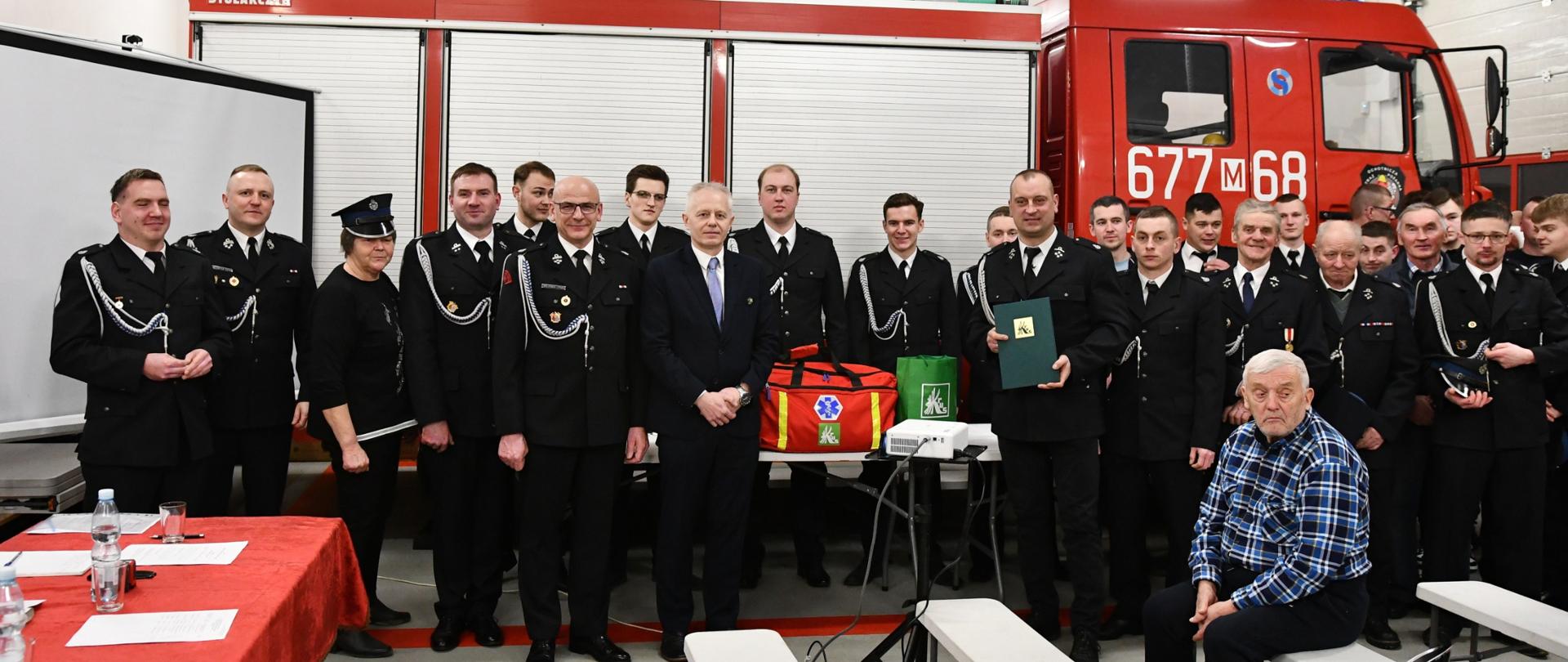Kobieta i kilkudziesięciu mężczyzn w galowych mundurach OSP oraz mężczyzna w cywilnym ubraniu pozują do zdjęcia z torbą ratownictwa przedmedycznego na tle bojowego wozu strażackiego. Przed wszystkimi na ławce siedzi starszy mężczyzna w niebieskiej koszuli w kratę.