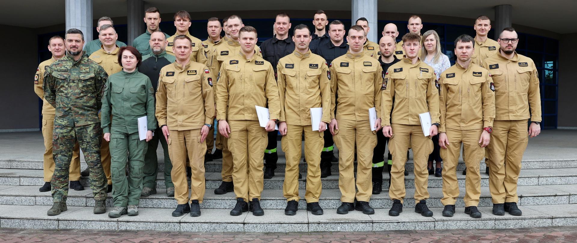 Pamiątkowe zdjęcie funkcjonariuszy PSP oraz Straży Granicznej (22 osoby) będących uczestnikami szkolenia w zakresie przeciwdziałania zagrożeniom chemicznym, biologicznym, radiacyjnym, nuklearnym i wybuchowym w towarzystwie Zastępcy Komendanta CS PSP, kobiety oraz trzech funkcjonariuszy CS PSP