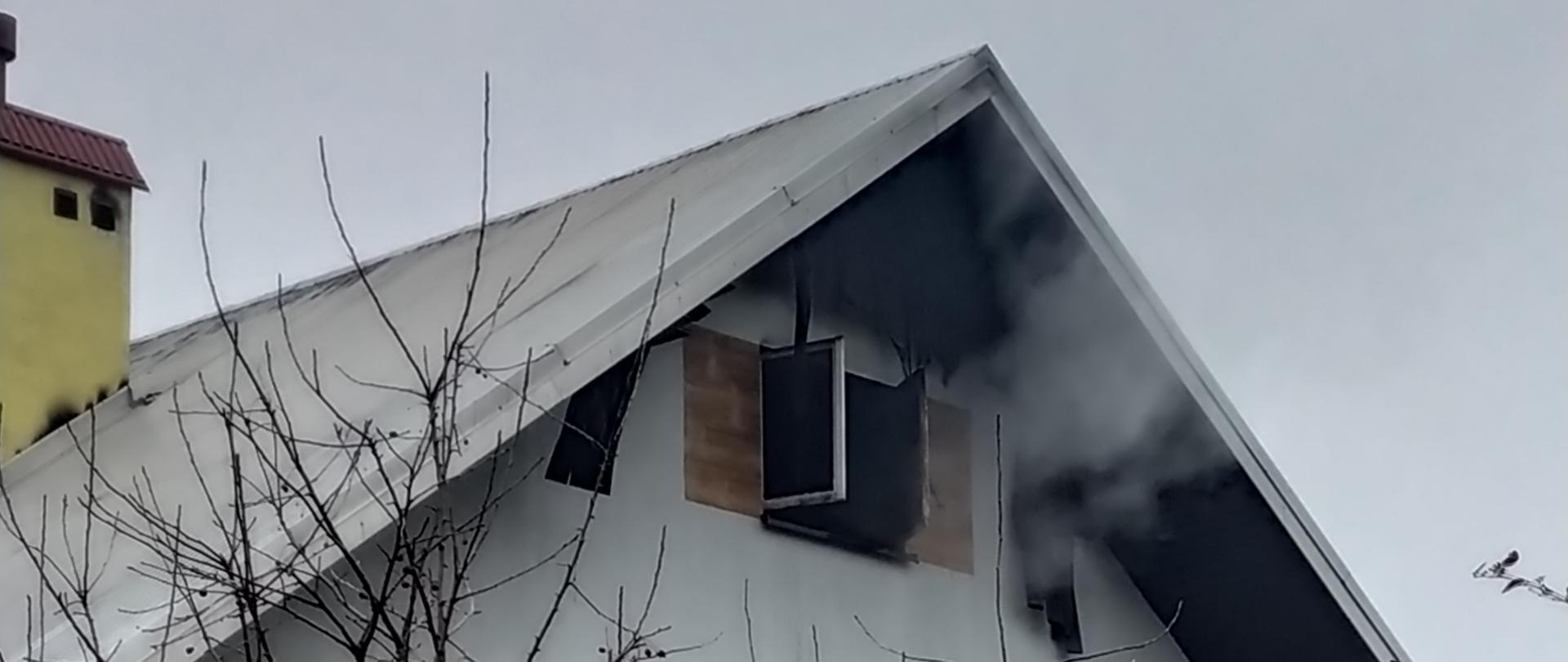 Na zdjęciu widać uchodzący z okna strychowego domu jednorodzinnego. Dom jest piętrowy i wykonany w konstrukcji murowanej. Przed domem rosną drzewa.