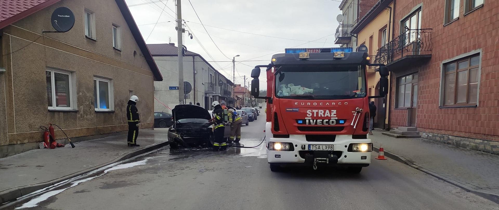 Zdjęcie przedstawia samochód osobowy, który gaszą strażacy, samochód stoi wzdłuż drogi przy krawężniku, po prawej stronie widać wóz strażacki z włączonymi lampami błyskowymi, po lewej widać budynek .
