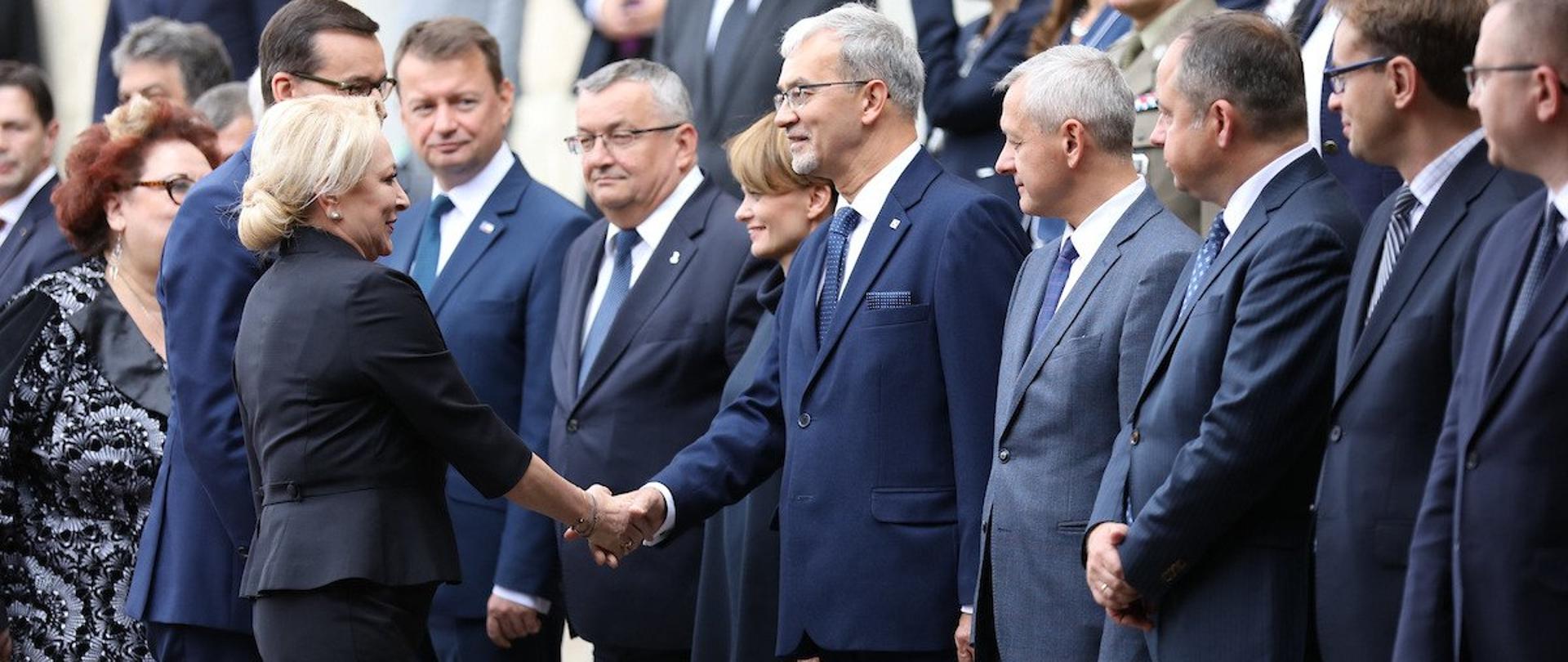 W rzędach stoi grupa ludzi. Witają się z nimi premierzy Polski i Rumunii. Minister Jerzy Kwieciński podaje rękę premier Rumunii.