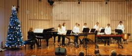Scena w kolorze berzowym-brązowym, na której występuje zespół perkusyjny w strojach biało czarnych. po prawej stoi choinka świąteczna, na fortepianie akompaniuje pani wice dyrektor Agnieszka Tyrała 