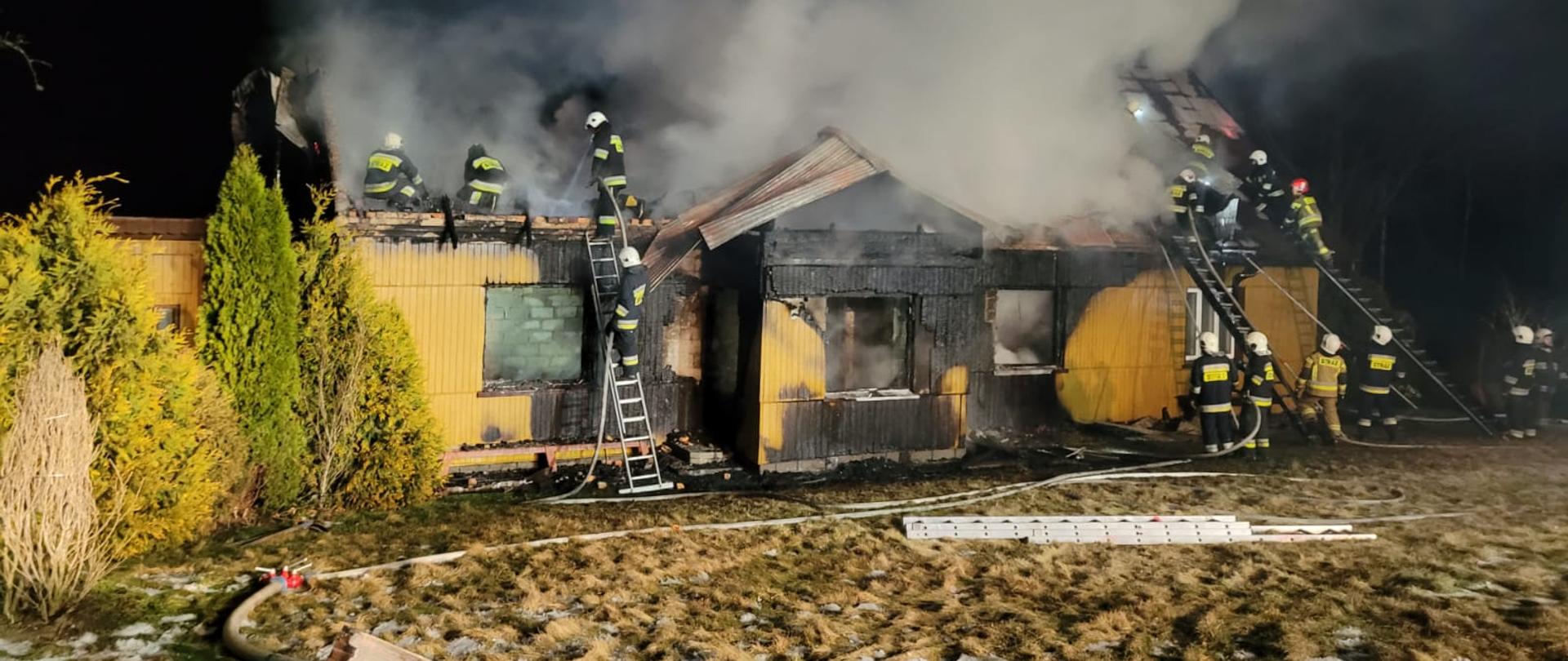 Zdjęcie przedstawia strażaków pracujących podczas pożaru budynku drewnianego. 