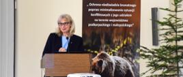Sala konferencyjna. Przed mównicą z mikrofonem stoi kobieta. Z tyłu plakat ze zdjęciem niedźwiedzia brunatnego i napisem: „Ochrona niedźwiedzia brunatnego poprzez minimalizowanie sytuacji konfliktowych z jego udziałem na terenie województwa podkarpackiego i małopolskiego”