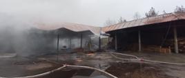 Zdjęcie przedstawia spalony budynek tartaku. W budynku unosi się dym. Przed budynkiem widać linie wężowe. 