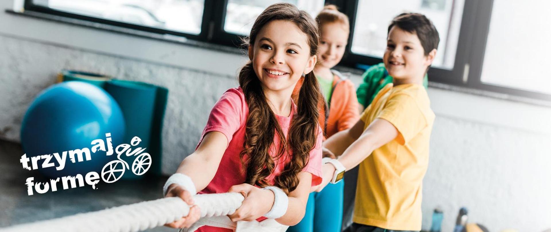 Zdjęcie przedstawia uśmiechnięte dzieci bawiące się w przeciąganie liny, w lewym dolnym rogu logotyp Programu "Trzymaj Formę!"