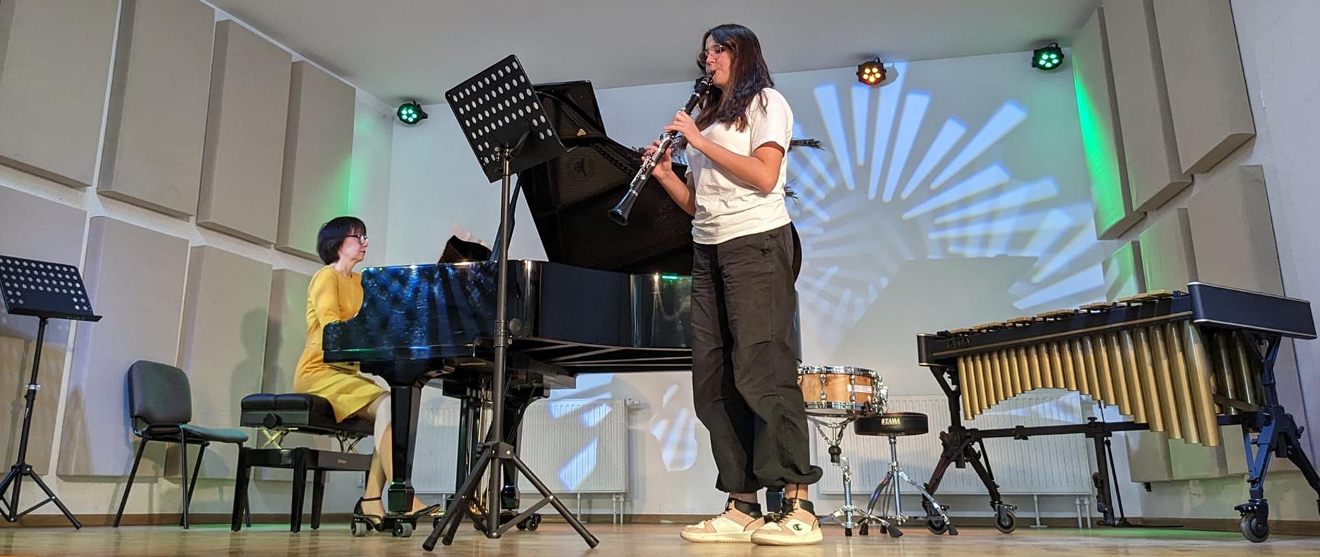 Klarnecistka wraz z akompaniatorką na scenie w sali koncertowej szkołu podczas koncertu. Z prawej strony stoi wibrafoan, a na tylnej ścianie mienią się kolorowe światła. 