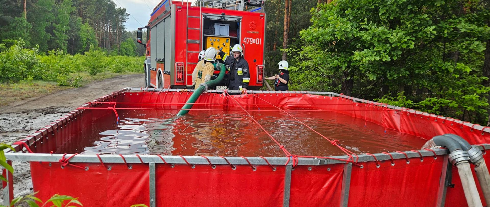 Czerwony zbiornik do magazynowania wody, częściowo wypełniony, stoi na ziemi. Do zbiornika włożony jest wąż. W tle widoczny jest czerwony wóz strażacki i strażacy. Całość znajduje się w lesie.