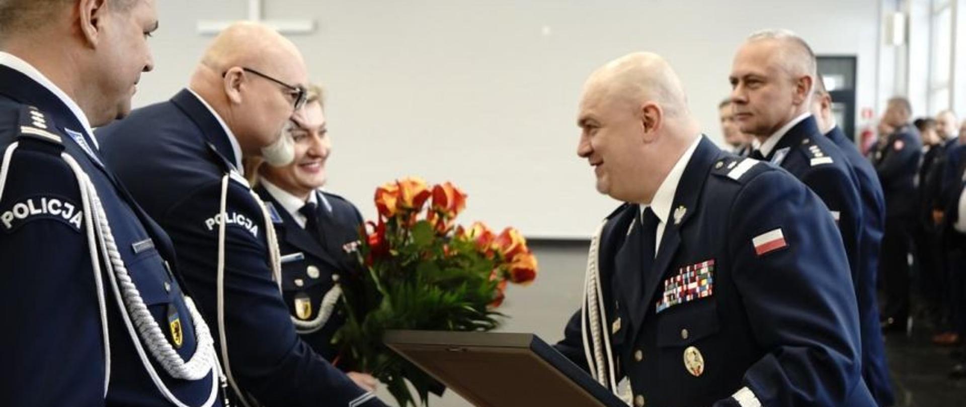 Czterech mężczyzn i kobieta (policjanci). Jeden mężczyzna podaje rękę drugiemu mężczyźnie i wręcza mu obraz. Kobieta trzyma bukiet kwiatów (róże).